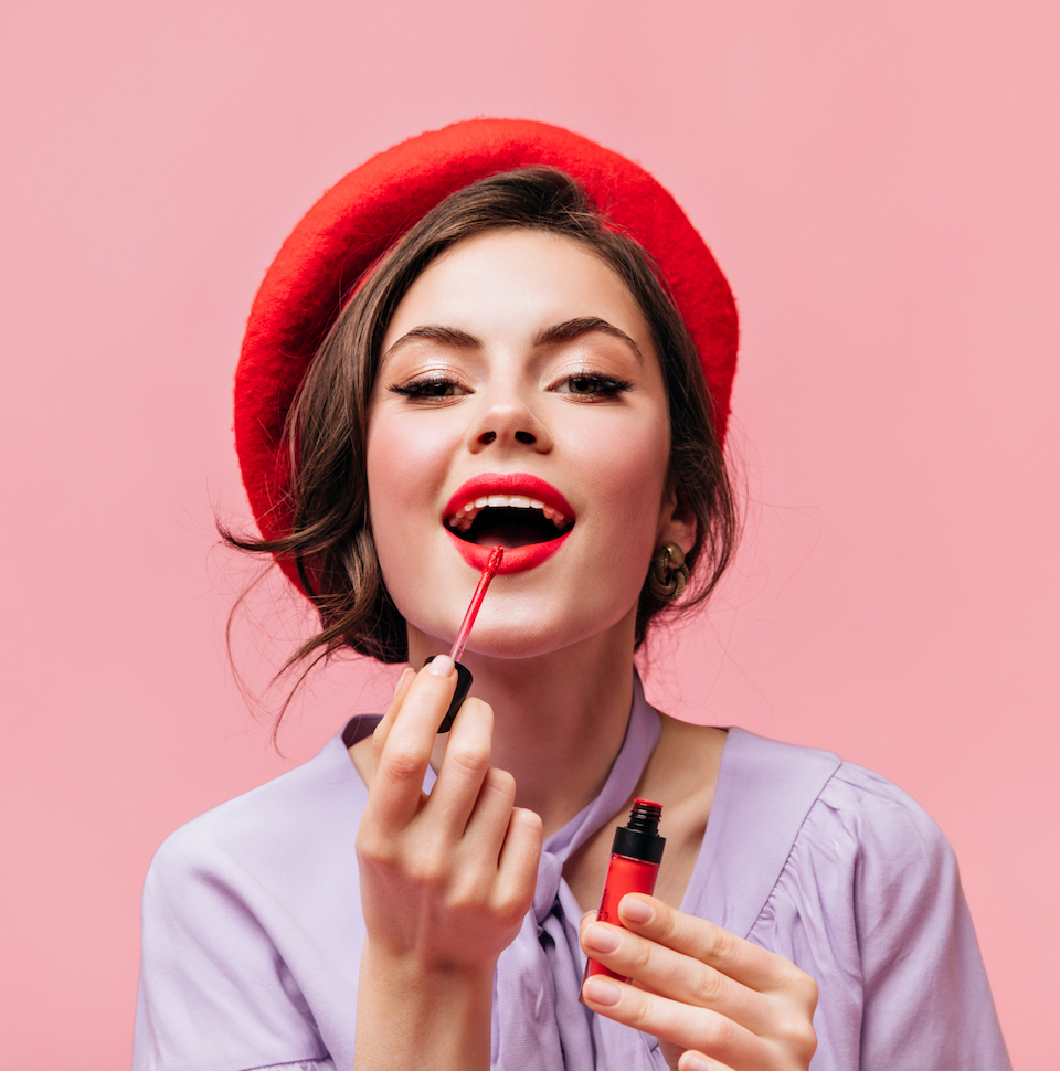 Woman in bright beret paints her lips with red lipstick. Girl in lilac blouse posing on pink background.