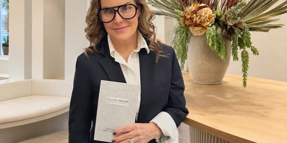 Christine Morrison holding her book Clothes Minded while wearing a black blazer and white shirt in a minimalist interior setting.