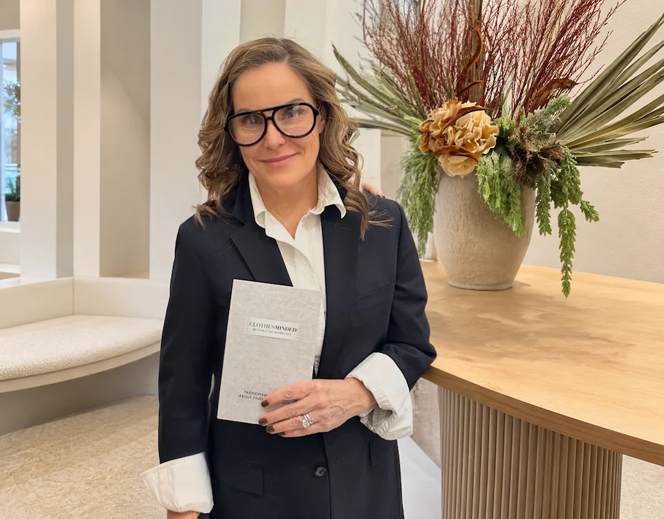 Christine Morrison holding her book Clothes Minded while wearing a black blazer and white shirt in a minimalist interior setting.