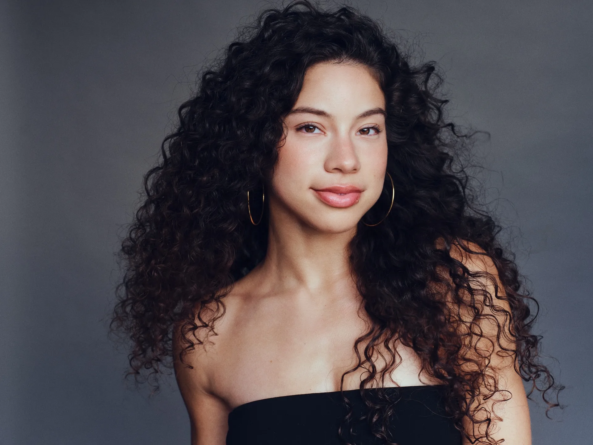 Actress Kiara Romero with long curly hair wearing a black strapless top in an editorial portrait.