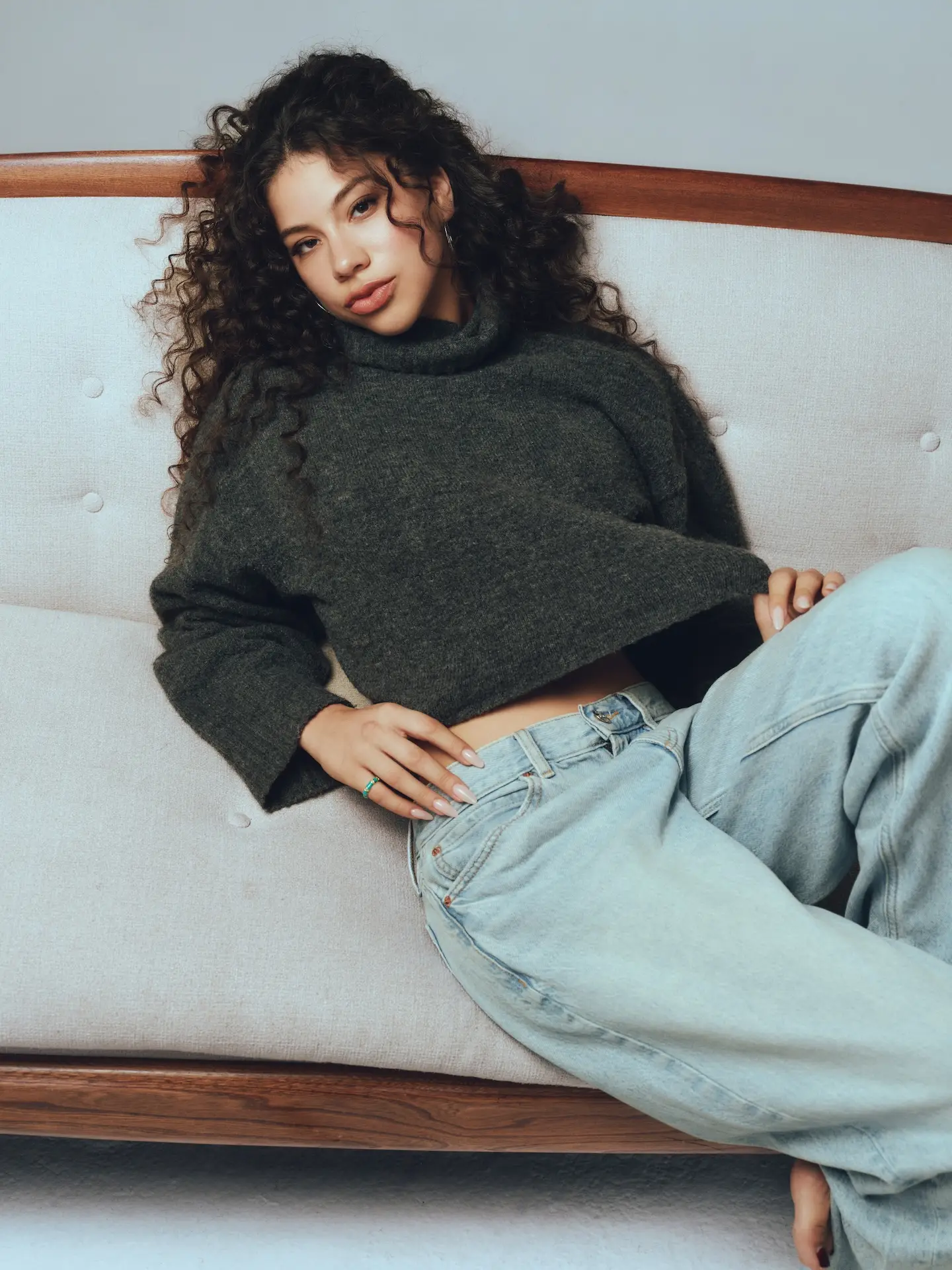Actress Kiara Romero sitting upright and reclining against a couch in an editorial portrait.
